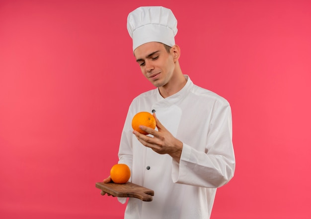 Jovem cozinheiro satisfeito vestindo uniforme de chef segurando e olhando laranja na tábua com espaço de cópia