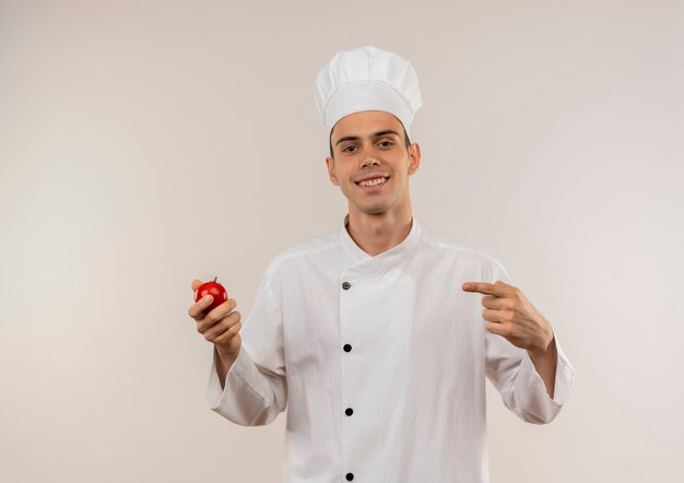 Jovem cozinheiro sorridente usando uniforme de chef apontando o dedo para o tomate na mão