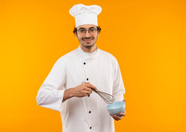 Jovem cozinheiro sorridente usando uniforme de chef e óculos segurando um batedor e uma tigela isolados na parede amarela