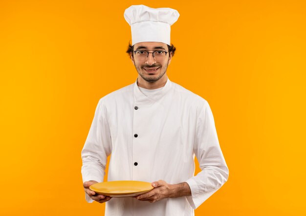 Jovem cozinheiro sorridente usando uniforme de chef e óculos segurando um prato isolado na parede amarela