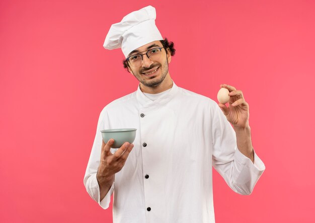 Jovem cozinheiro sorridente usando uniforme de chef e óculos segurando uma tigela e um ovo isolado na parede rosa