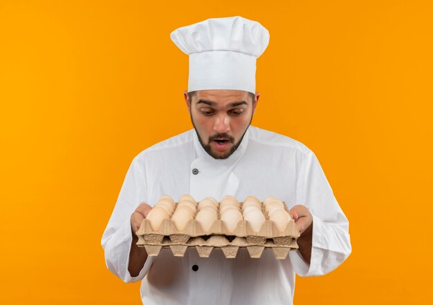 Foto grátis jovem cozinheiro surpreso com uniforme de chef segurando e olhando para uma caixa de ovos isolada no espaço laranja