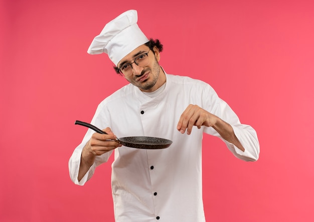 Jovem cozinheiro usando uniforme de chef e óculos, segurando uma frigideira e fingindo derramar sal