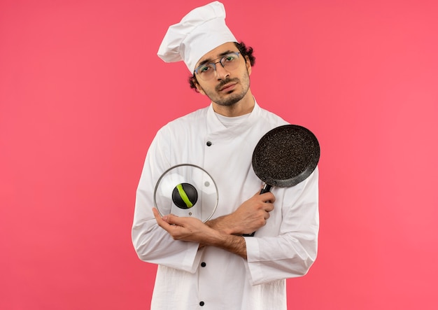 Jovem cozinheiro vestindo uniforme de chef e óculos segurando e cruzando a frigideira com tampa