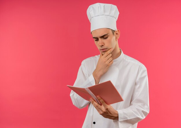 Jovem cozinheiro vestindo uniforme de chef segurando e olhando para noyebook colocando a mão no queixo com espaço de cópia