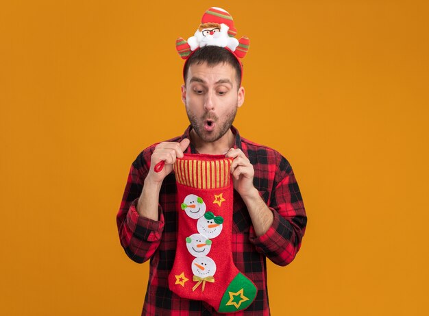 Jovem curioso, caucasiano, usando uma bandana de papai noel segurando uma meia de natal, abrindo e olhando dentro dela, isolado em uma parede laranja com espaço de cópia