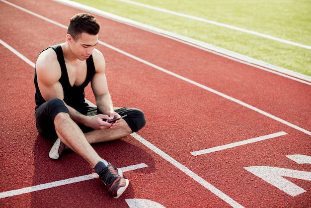 Jovem de aptidão usando telefone celular enquanto está sentado na pista de corrida