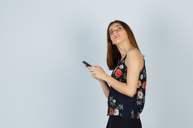 Foto grátis jovem de blusa segurando o smartphone enquanto olha para cima e parece pensativa.