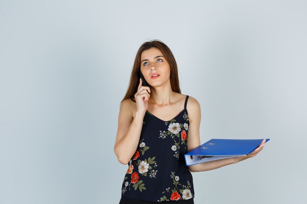 Jovem de blusa, segurando uma pasta, falando no celular e olhando pensativa, vista frontal.