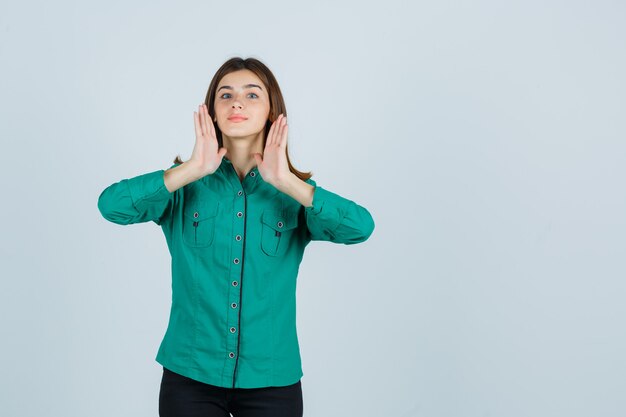 Jovem de blusa verde, calça preta de mãos dadas perto da cabeça e olhando bonita, vista frontal.