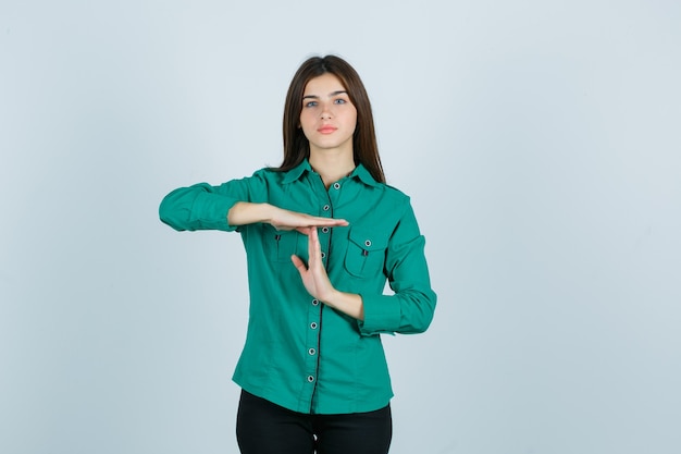 Jovem de blusa verde, calça preta, mostrando o gesto de pausa e olhando séria, vista frontal.