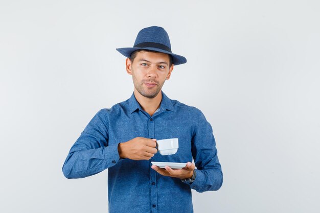 Jovem de camisa azul, chapéu, bebendo café turco e olhando positiva, vista frontal.