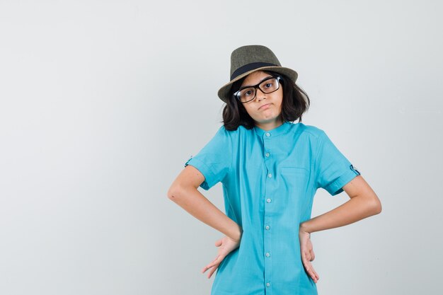 Foto grátis jovem de camisa azul, chapéu e óculos em pé com as mãos na cintura e parecendo triste