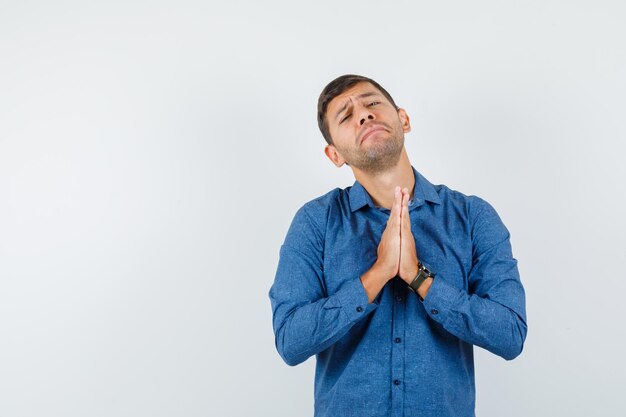 Jovem de camisa azul, de mãos dadas em gesto de oração e olhando impotente, vista frontal.