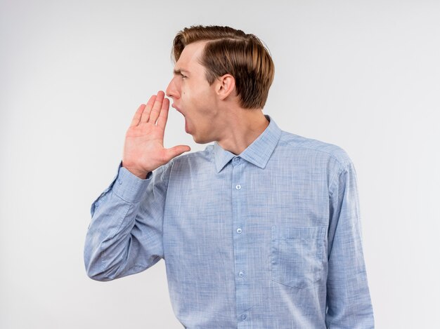 Jovem de camisa azul olhando para o lado gritando com a mão perto da boca, em pé sobre uma parede branca