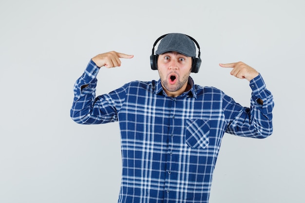 Jovem de camisa, boné, apontando para seus fones de ouvido e olhando surpreso, vista frontal.