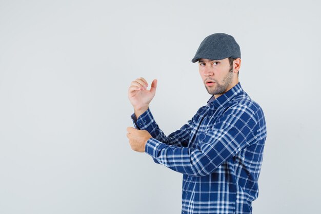 Jovem de camisa, boné desabotoando a manga da camisa e olhando pensativo, vista frontal.