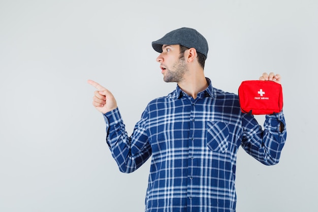 Jovem de camisa, boné segurando o kit de primeiros socorros, apontando para cima e olhando com foco, vista frontal.