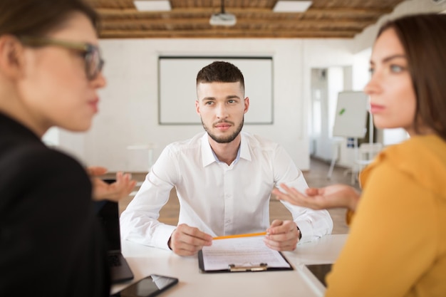 Jovem de camisa branca olhando pensativamente para os empregadores enquanto passa o tempo no escritório moderno Candidato masculino segurando lápis esperando resultados da entrevista de emprego