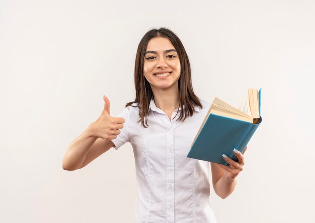 Jovem de camisa branca segurando um livro, olhando para a frente, mostrando os polegares em pé sobre uma parede branca