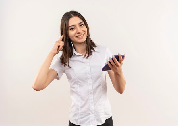 Jovem de camisa branca segurando um smartphone, apontando para a têmpora, parecendo confiante em pé sobre uma parede branca