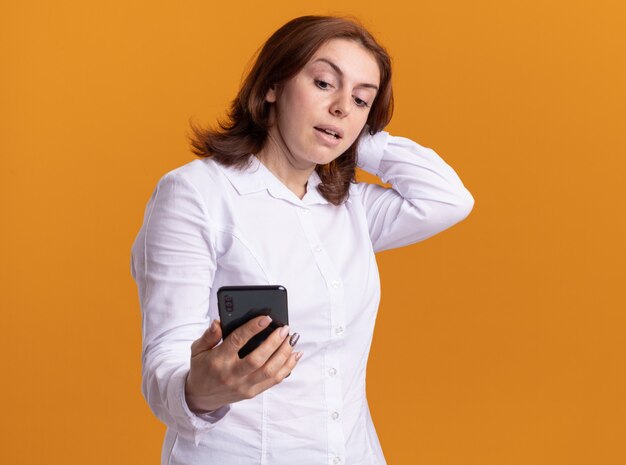 Jovem de camisa branca segurando um smartphone, olhando para ele sendo confusa em pé sobre uma parede laranja