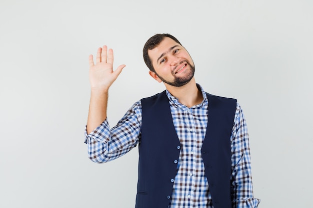 Jovem de camisa, colete acenando a mão para saudação e olhando alegre, vista frontal.