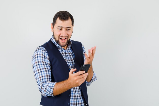 Jovem de camisa, colete, olhando para o celular e olhando alegre, vista frontal.