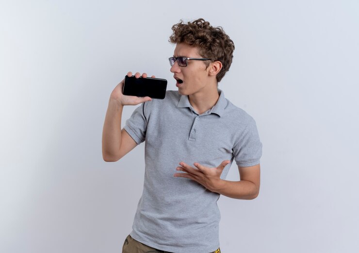Foto grátis jovem de camisa pólo cinza segurando um smartphone e usando como microfone cantando em pé sobre uma parede branca