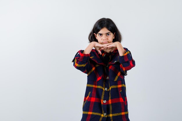 Jovem de camisa quadrada, colocando as mãos sob o queixo e olhando fofa, vista frontal.