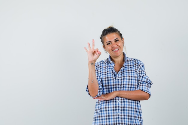 Jovem de camisa quadriculada, mostrando o gesto de ok e olhando alegre.