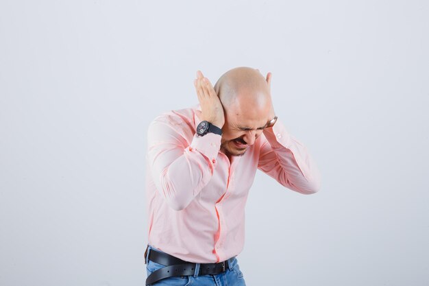 Jovem de camisa rosa, jeans de mãos dadas nas orelhas enquanto se inclina para a frente e parece louco, vista frontal.