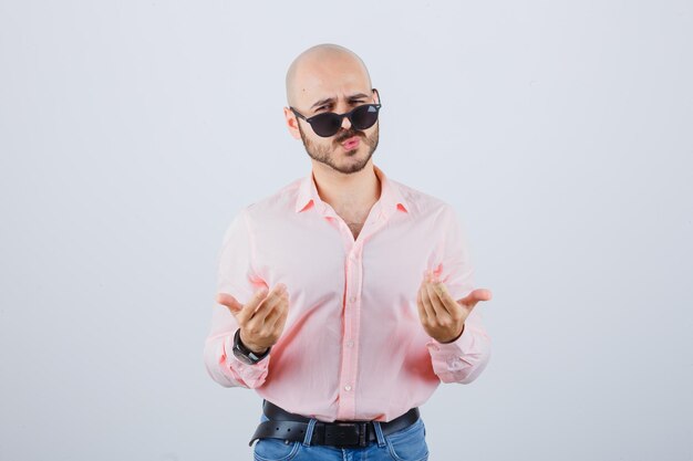 Jovem de camisa rosa, jeans, óculos de sol, expressando seus sentimentos com gestos com as mãos e parecendo estranho, vista frontal.