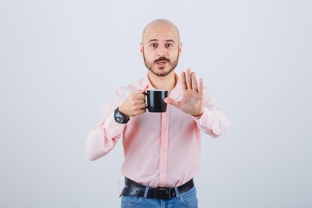 Jovem de camisa rosa, jeans, segurando o copo enquanto mostra o gesto de pare e parecendo alarmado, vista frontal.