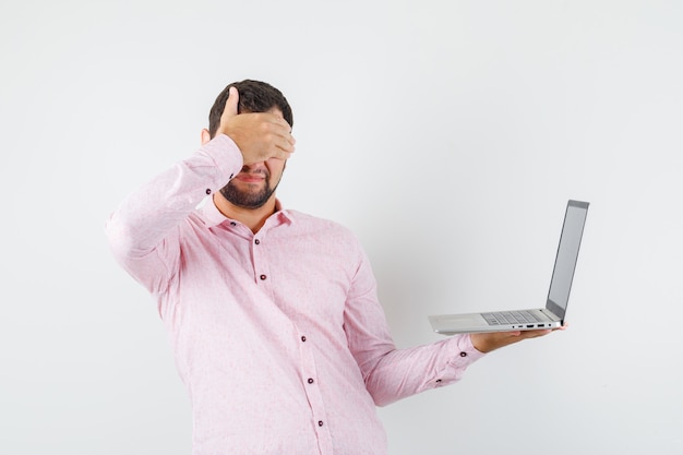 Jovem de camisa rosa segurando um laptop com a mão nos olhos