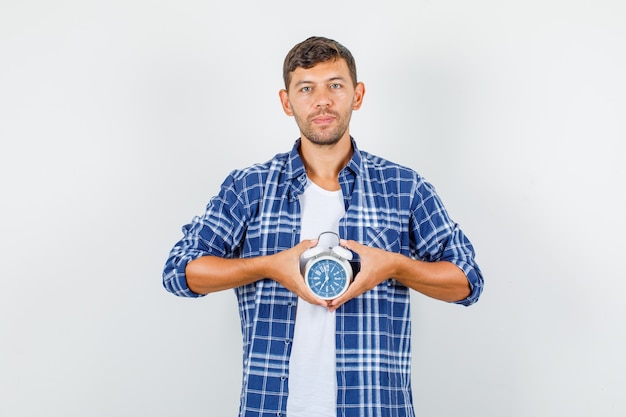 Jovem de camisa segurando o despertador e olhando vista frontal pontual.