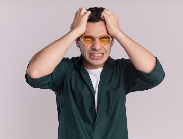 Jovem de camisa verde usando óculos enlouquecendo puxando o cabelo em pé sobre uma parede branca