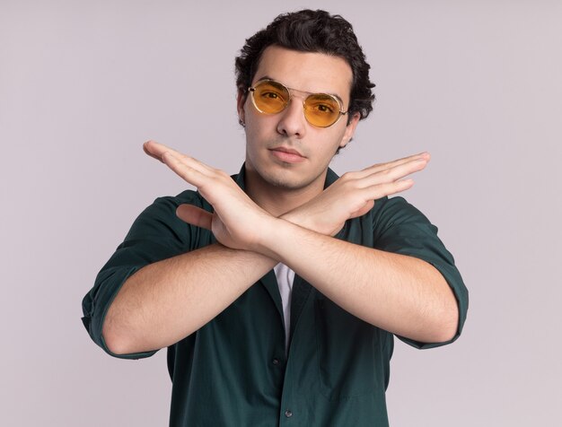 Jovem de camisa verde usando óculos, olhando para a frente com uma cara séria, cruzando as mãos, parando o gesto em pé sobre uma parede branca