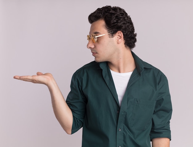 Jovem de camisa verde usando óculos, olhando para o lado, apresentando com o braço da mão copiar o espaço em pé sobre uma parede branca