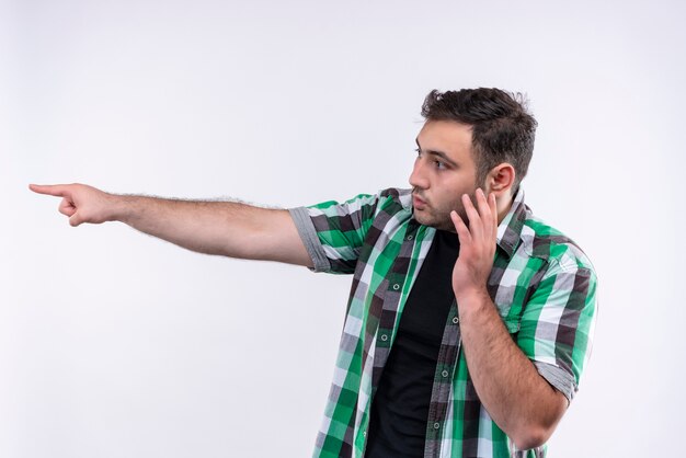 Jovem de camisa xadrez apontando com o dedo para o lado e parecendo confuso enquanto fala ao telefone celular em pé sobre uma parede branca