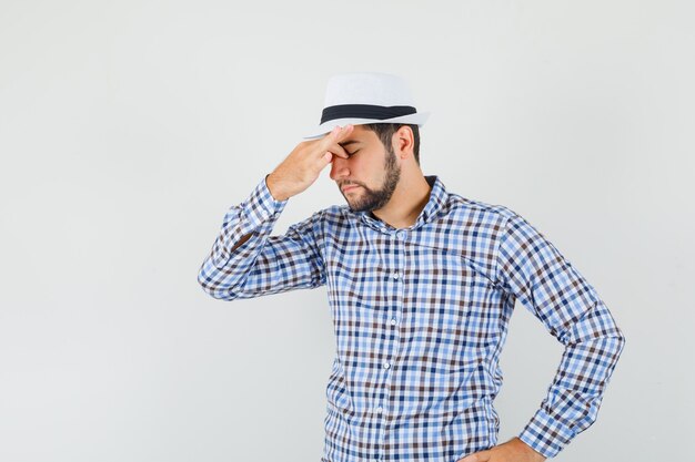 Jovem de camisa xadrez, chapéu esfregando os olhos e o nariz e parecendo fatigado, vista frontal.