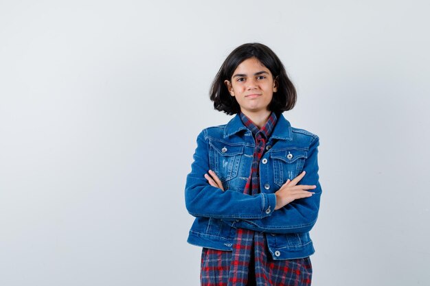 Jovem de camisa xadrez e jaqueta jeans em pé com os braços cruzados e olhando feliz, vista frontal.