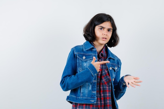 Jovem de camisa xadrez e jaqueta jeans, esticando uma mão, segurando algo e apontando para ele com o dedo indicador e olhando sério, vista frontal.