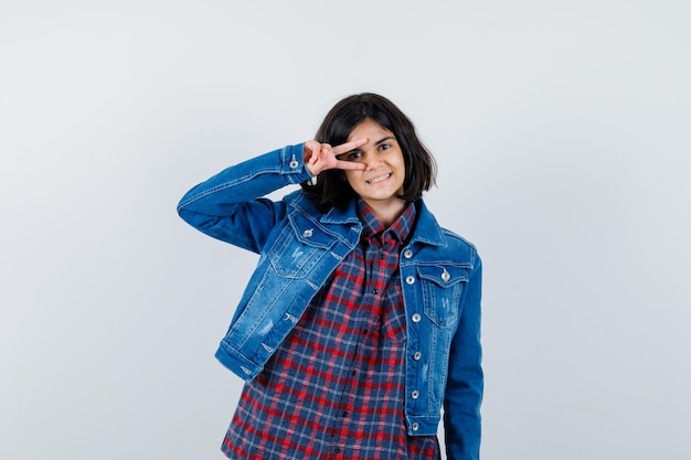 Jovem de camisa xadrez e jaqueta jeans, mostrando o sinal v no olho e está linda, vista frontal.