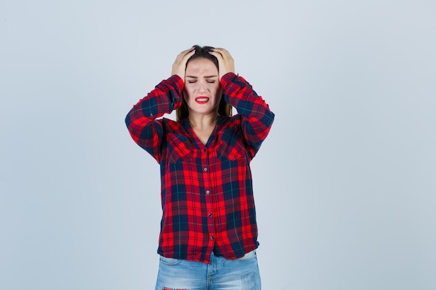 Jovem de camisa xadrez, jeans com as mãos na cabeça e olhando preocupada, vista frontal.