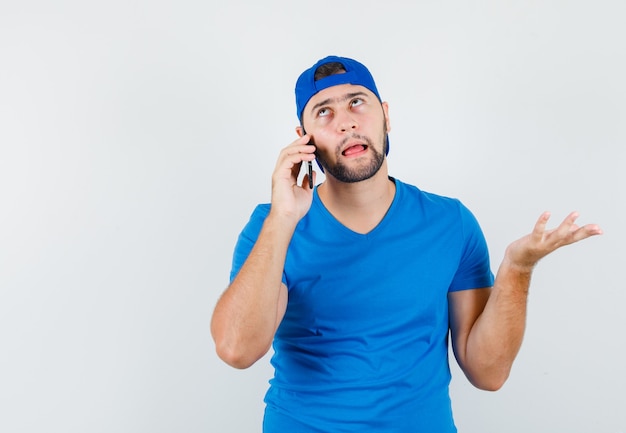 Jovem de camiseta azul e boné falando no celular e parecendo hesitante