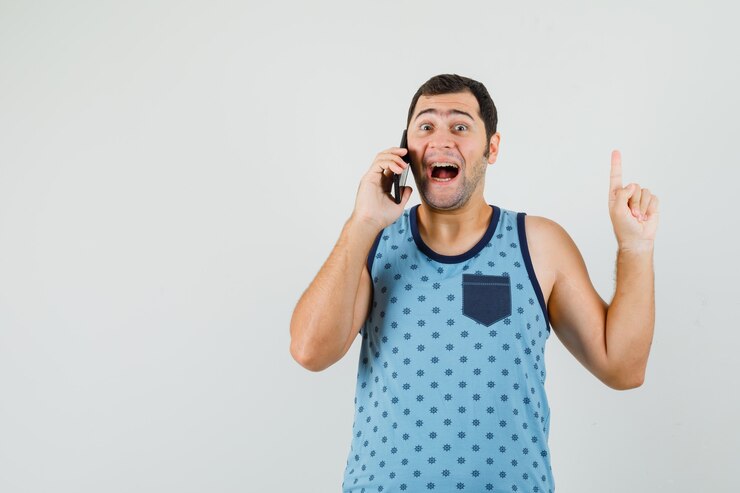 Foto grátis jovem de camiseta azul falando no celular, apontando para cima e parecendo feliz