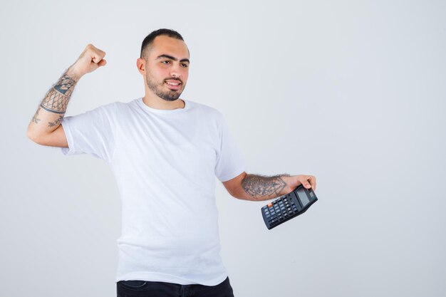 Jovem de camiseta branca e calça preta segurando uma calculadora e mostrando o gesto do vencedor e parecendo feliz