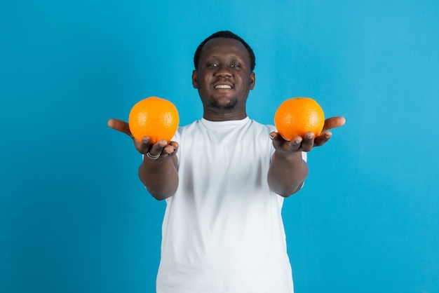 Jovem de camiseta branca segurando duas frutas de laranja doce contra uma parede azul