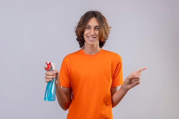 Foto grátis jovem de camiseta laranja segurando spray de limpeza e sorrindo alegremente olhando para a câmera apontando para o lado em pé sobre fundo branco
